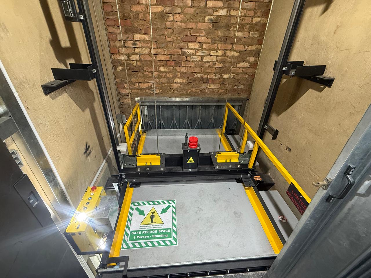 Goods hoist car top with yellow safety railings, a red buffer, marked safe refuge space decal and a limited-headroom warning sign in a brick shaft.