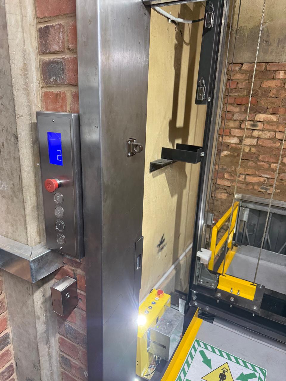 Stainless steel landing call panel with a blue digital position indicator, red stop button and four floor buttons, mounted beside the open hoist shaft.