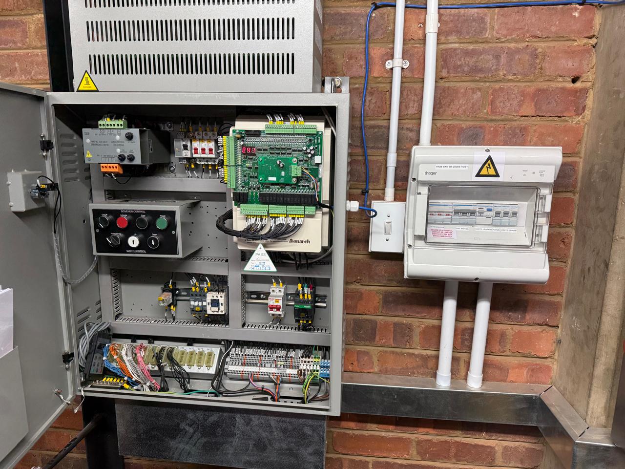 Open goods hoist control cabinet showing a Monarch controller board, maintenance control panel with governor controls, contactors and terminated wiring, next to a Hager distribution board.