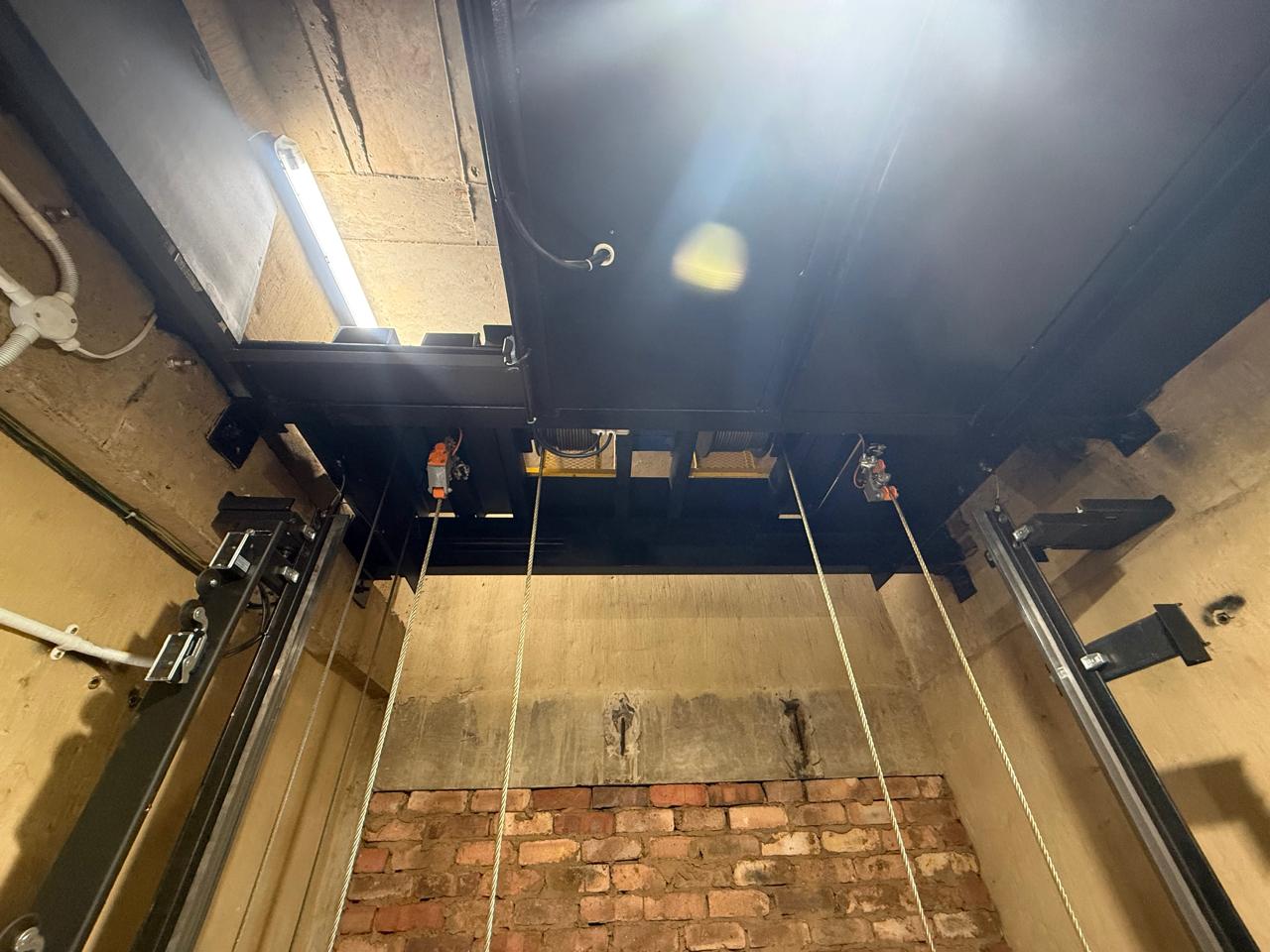 View up a goods hoist shaft to the underside of the car sling, showing hoisting ropes, guide rails, shaft lighting and safety gear.