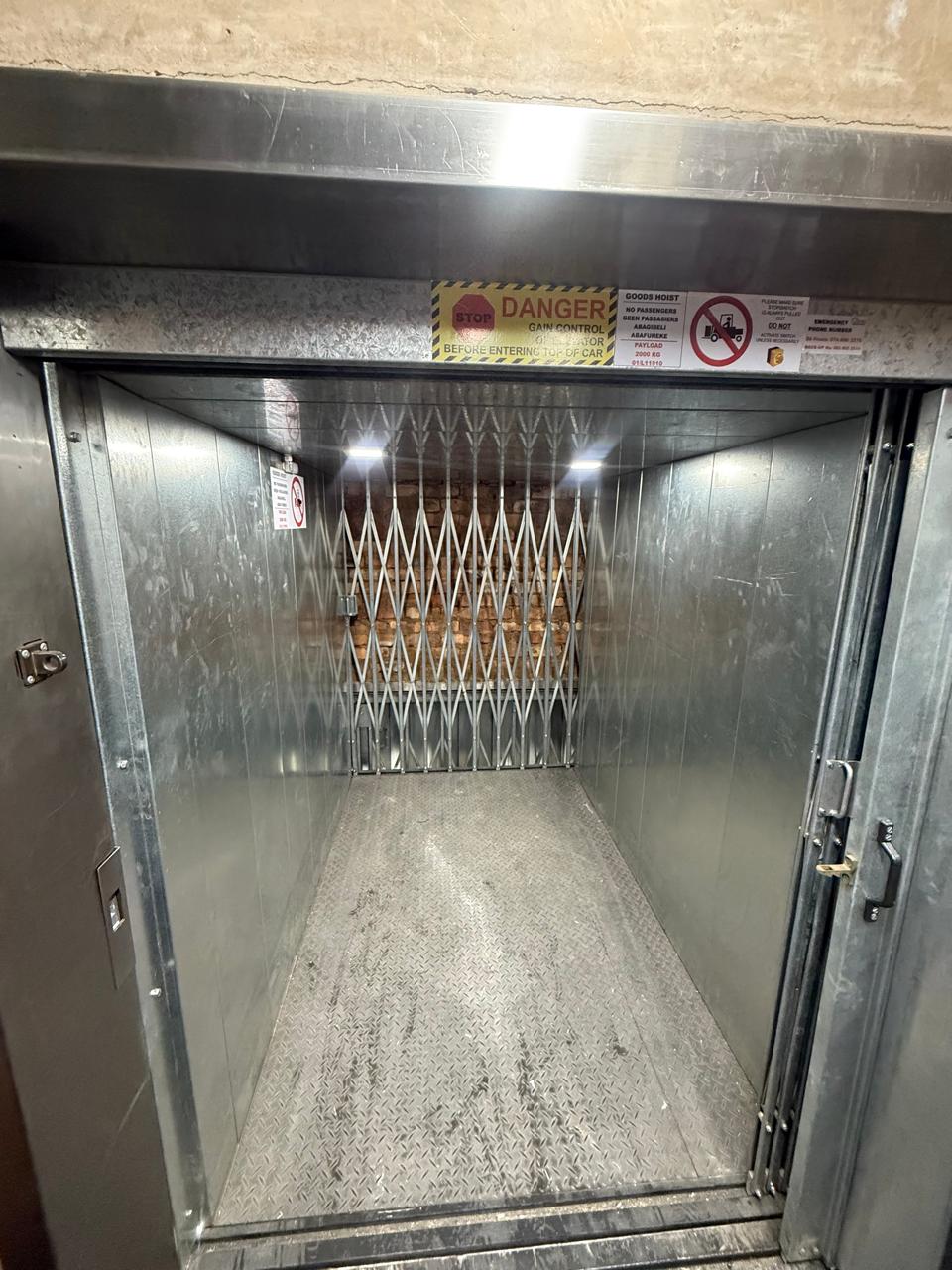 Galvanised goods hoist car interior with a concertina gate, chequer plate floor, LED lighting and a 2000 kg payload notice with no-passenger signage.