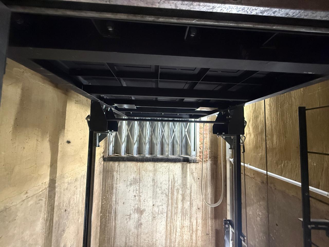 Underside of the goods hoist car showing the fabricated steel platform frame, safety gear blocks on the guide rails and a concertina gate at the landing beyond.