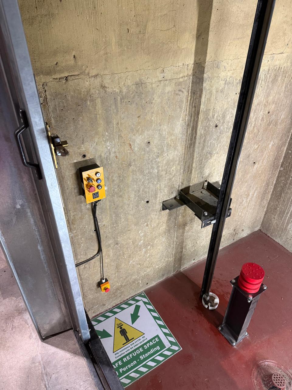 Goods hoist pit access with a yellow pit inspection control station, pendant stop switch, buffer on the sealed floor and a safe refuge space decal.