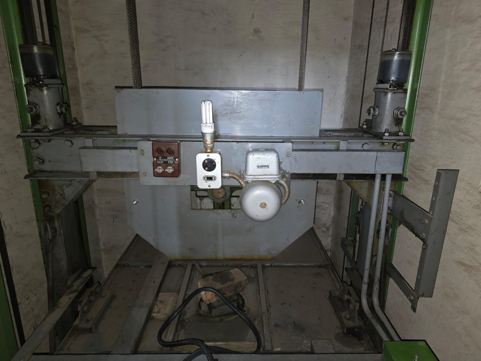 Original lift car top with no balustrade or refuge marking, an old inspection button station, alarm bell and a bare lamp.