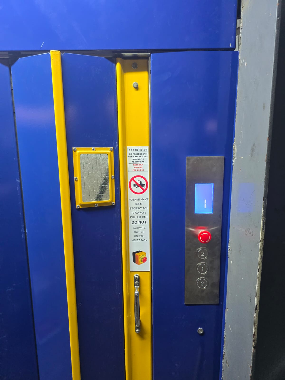 Blue folding goods hoist landing door with yellow trim, wired glass vision panel, 1500 kg payload notice and a call panel with position indicator.
