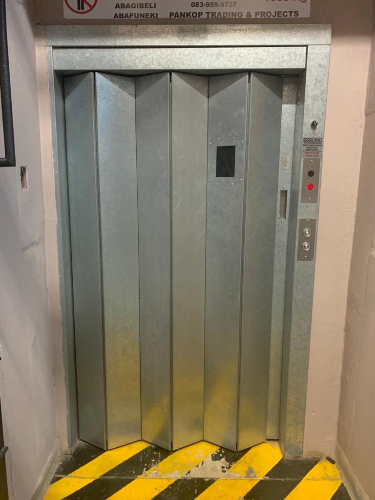 Galvanised folding goods hoist landing door with a call panel, no-passengers payload notice and hazard-striped threshold.
