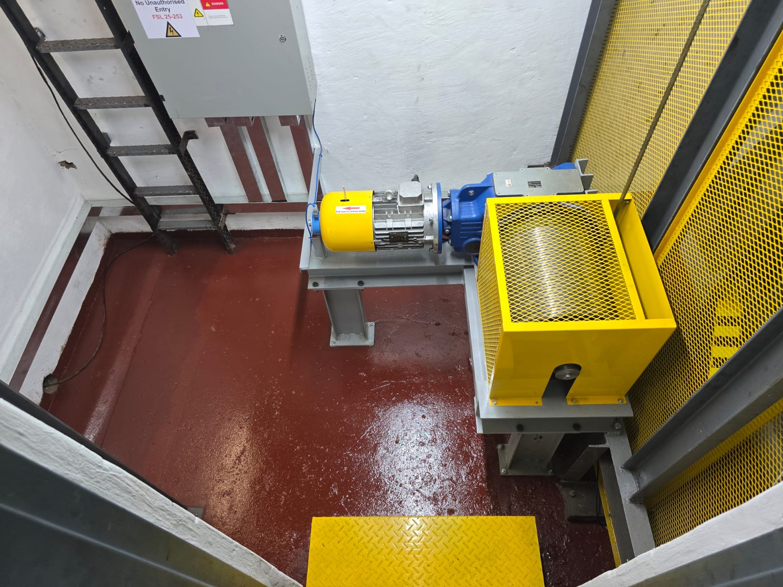 Goods hoist machine room with a yellow motor and blue gearbox on a fabricated bedplate, yellow mesh sheave guard, control cabinet and sealed floor.