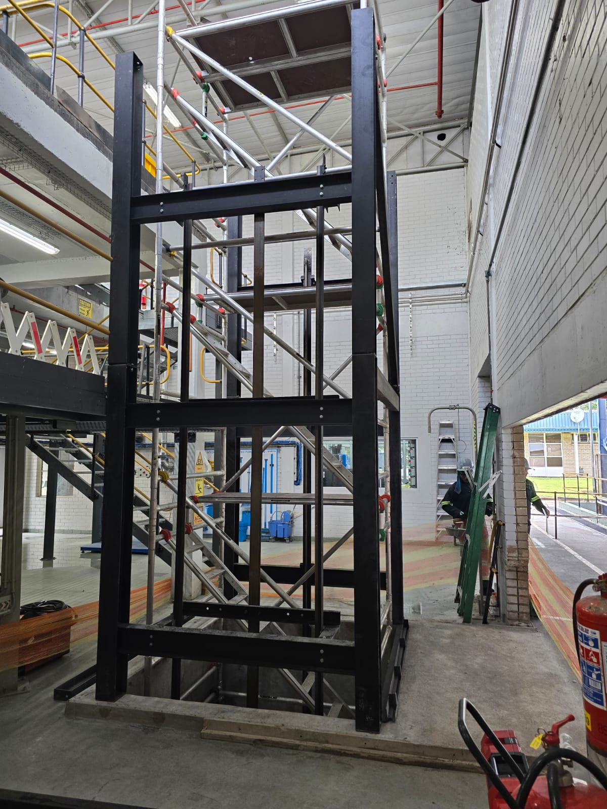 Fabricated black steel goods hoist shaft structure erected inside a working factory, with scaffolding around it during installation.