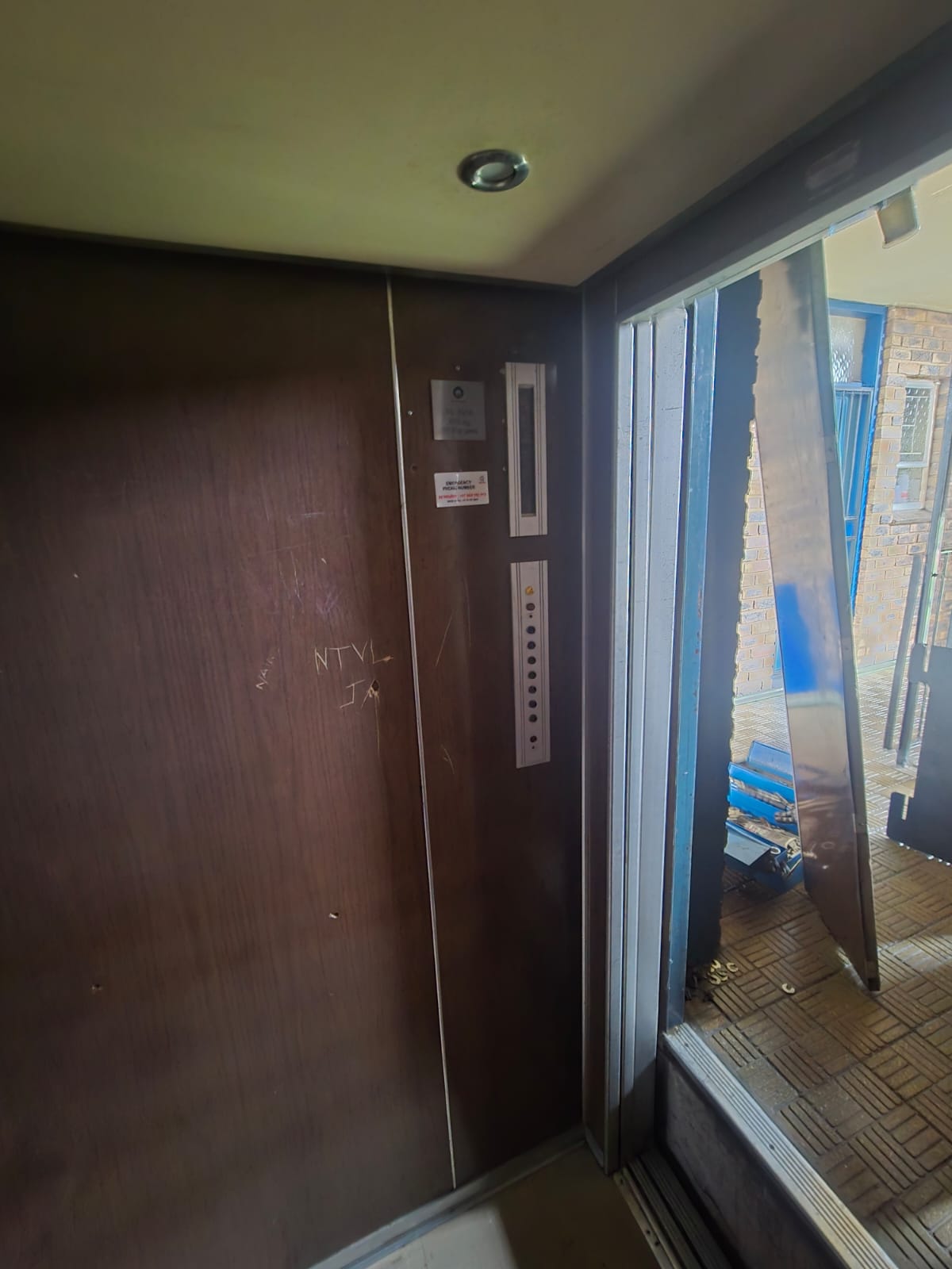 Lift cab due for refurbishment with scratched dark timber panels and a dated car operating panel, new stainless panels waiting outside the open door.