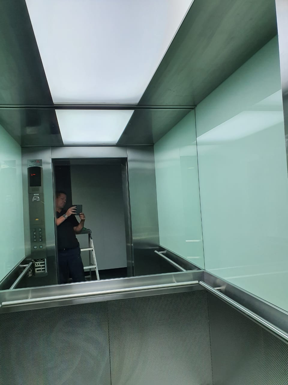 Lift cab with pale green back-painted glass upper panels, woven stainless lower panels and a large illuminated translucent ceiling.