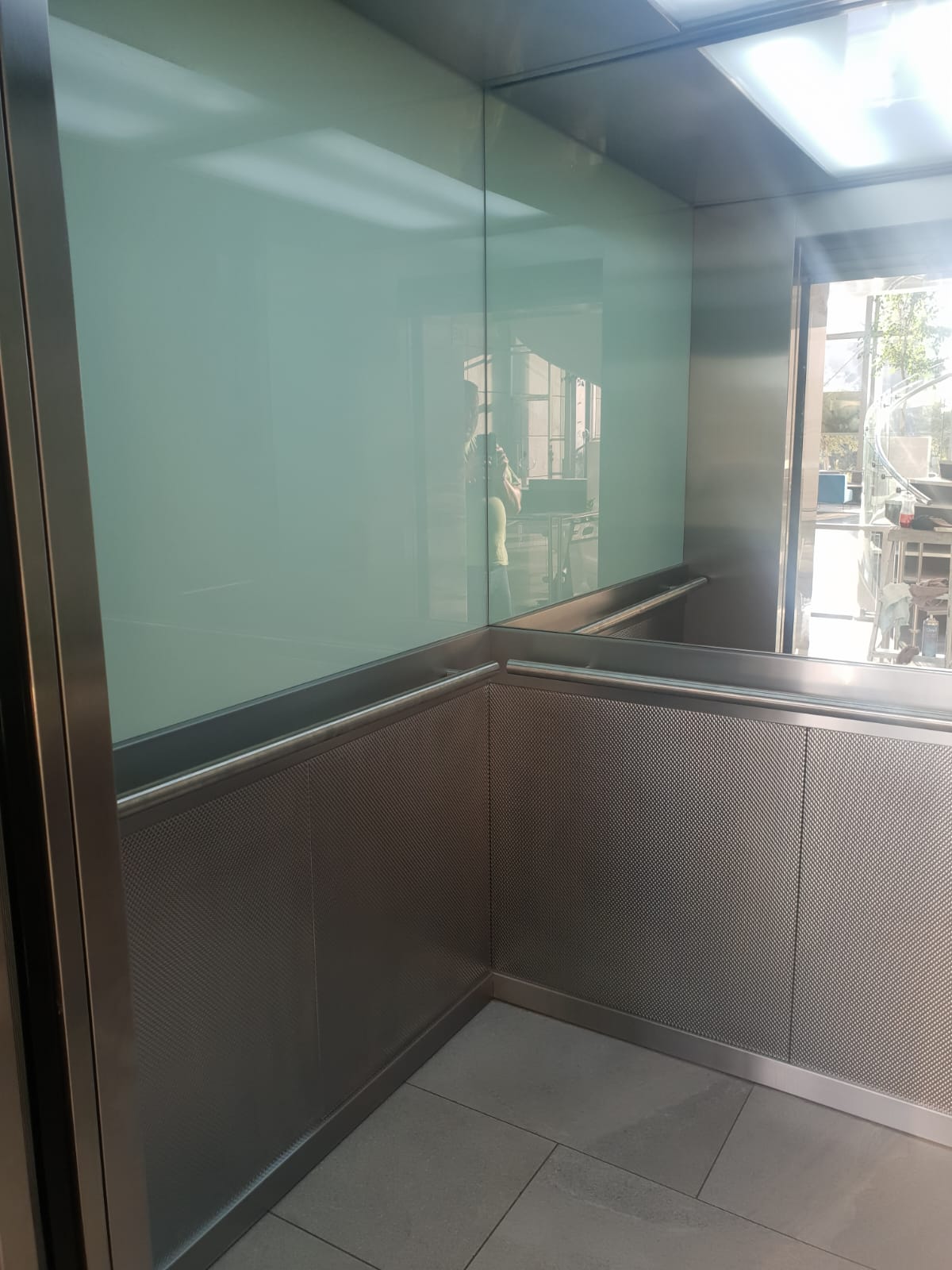 Lift cab corner showing pale green back-painted glass upper panels, woven-texture stainless lower panels and stainless handrail.