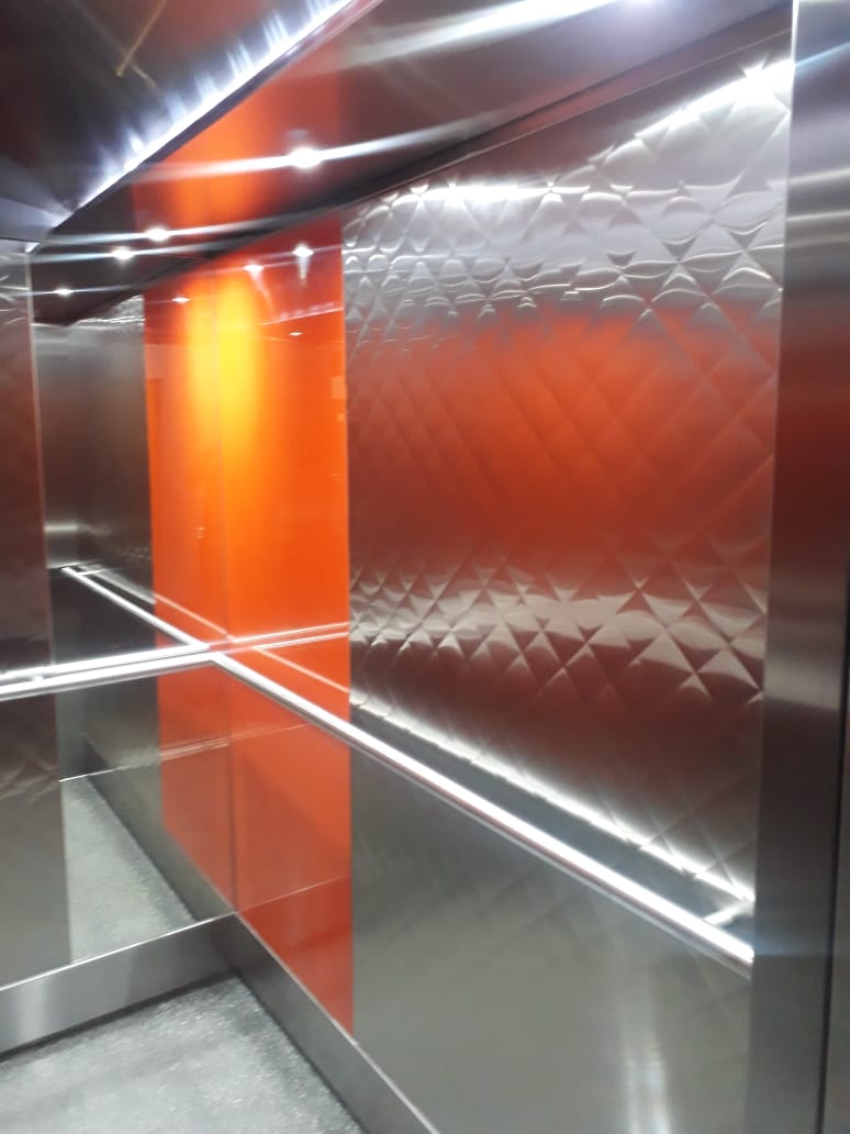 Lift interior detail showing embossed diamond-pattern stainless panels beside a gloss orange accent panel and stainless handrail.