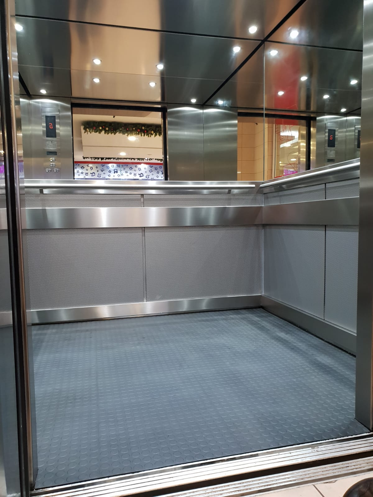 Large lift cab with woven-texture stainless lower panels, mirrored upper walls and ceiling with downlights, handrails on three sides and studded rubber flooring.
