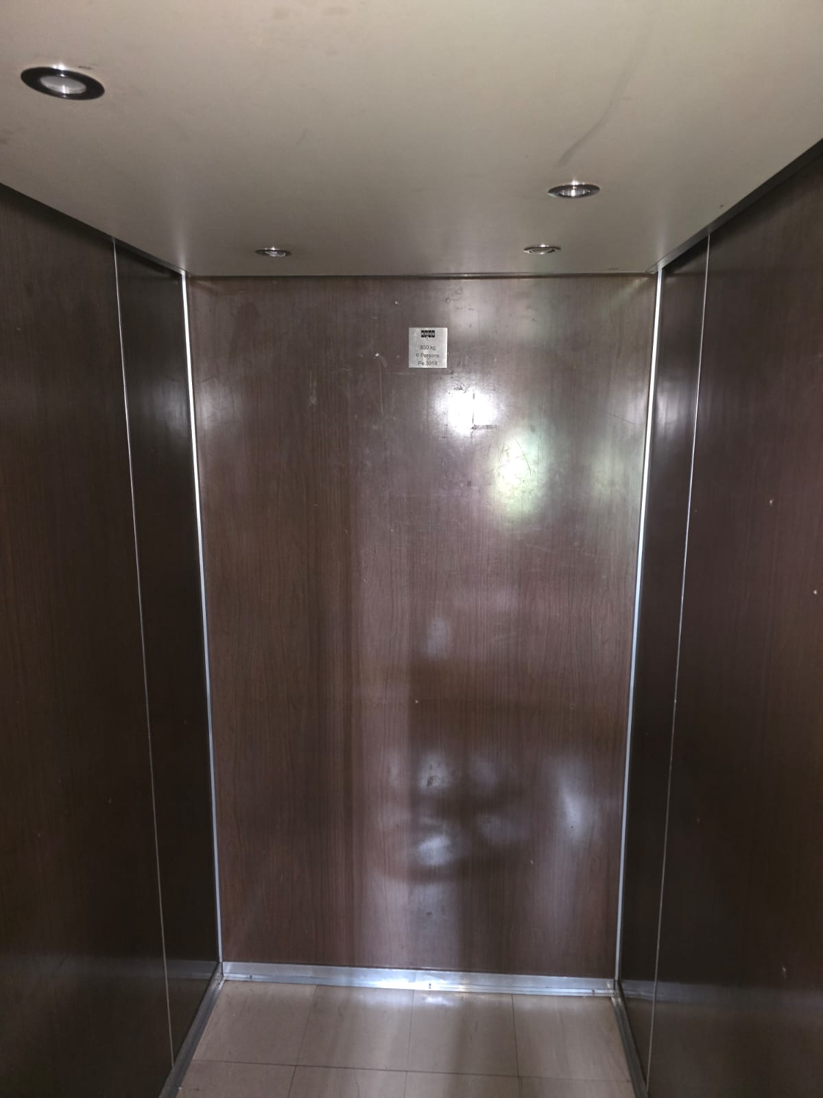 Lift cab with timber-look wall panels, aluminium trim, ceiling downlights, capacity plate and tiled flooring.