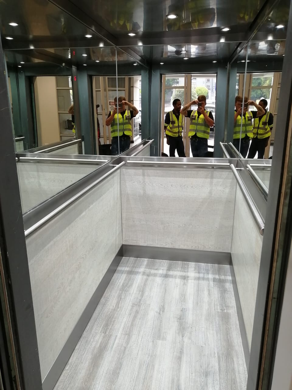Lift cab with pale timber-look wall panels, full mirror above the handrail, stainless handrails and grey plank flooring.