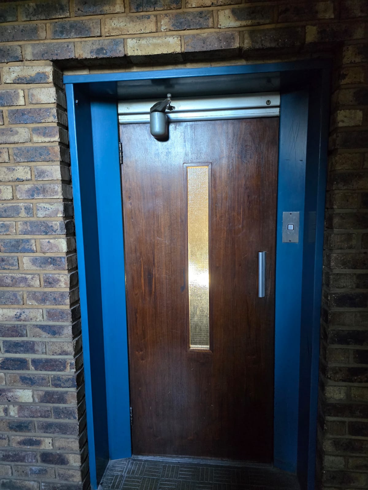 Old scratched timber swing landing door with a narrow glass vision strip, overhead closer and a single call button, set in a blue-painted frame.