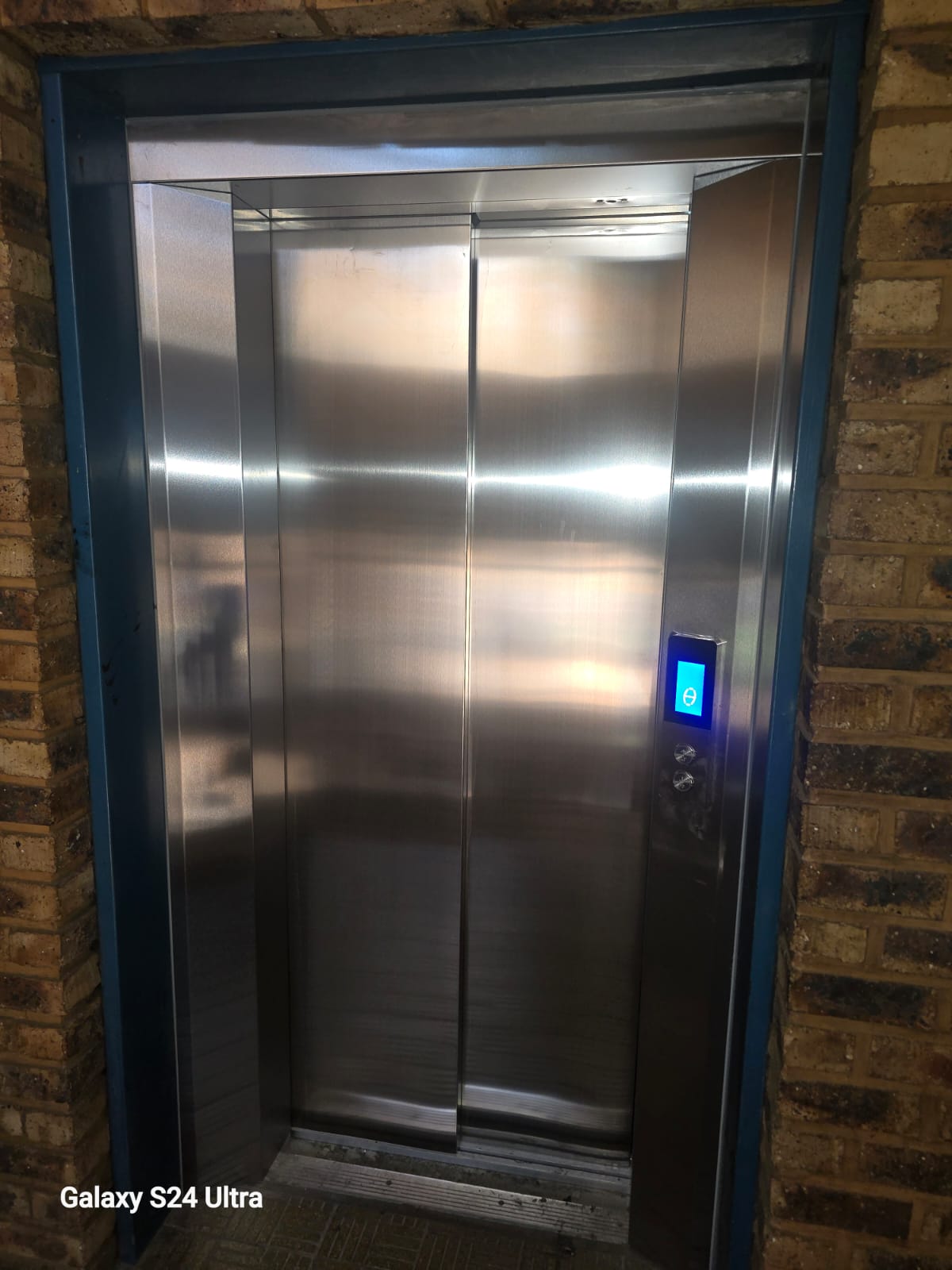 The same landing fitted with brushed stainless steel sliding doors, illuminated position indicator and call buttons, in the same blue-painted frame.