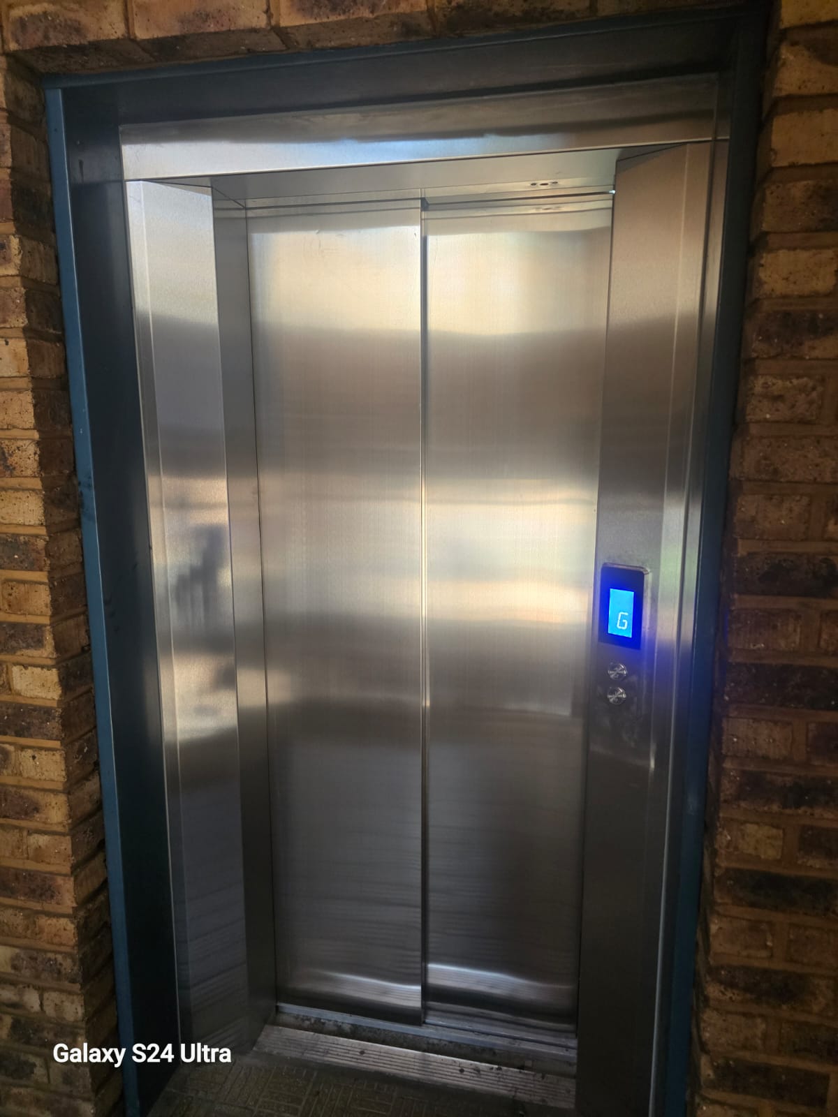 Brushed stainless steel landing doors and frame set into face brick, with an illuminated ground floor indicator and call buttons.