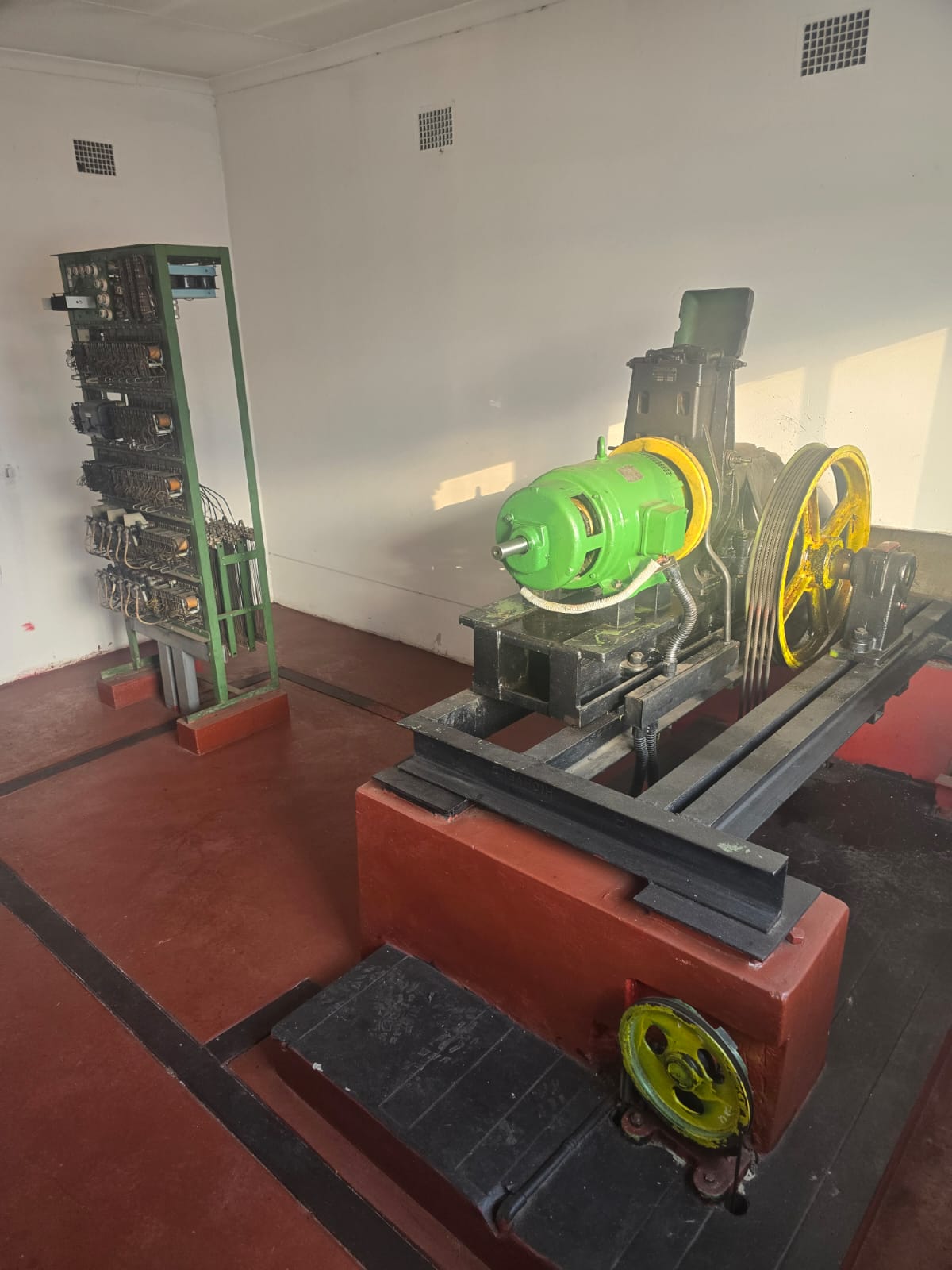 Machine room with a green open relay control frame, geared traction machine with yellow sheave, and diverter pulley set into a red plinth.