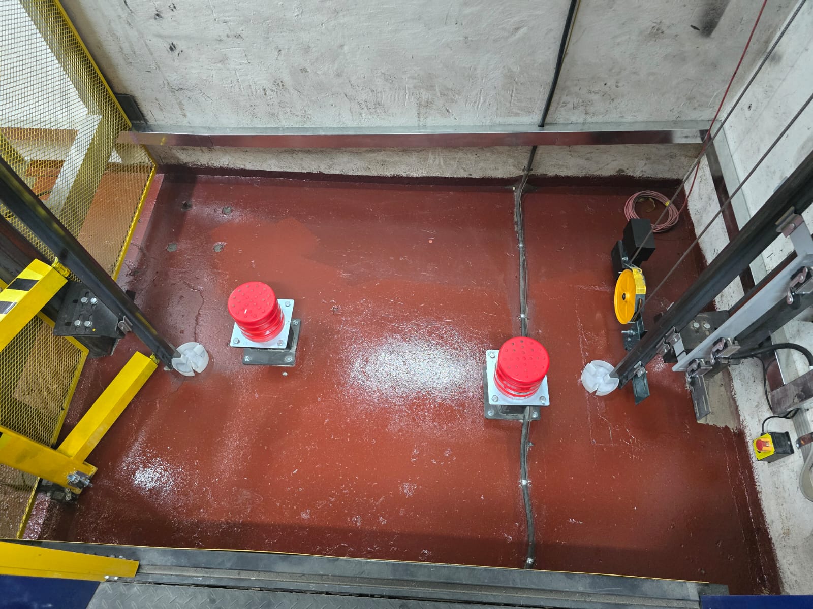 Sealed lift pit with two red buffers on mounting plates, guide shoes, governor tension sheave and conduit runs.